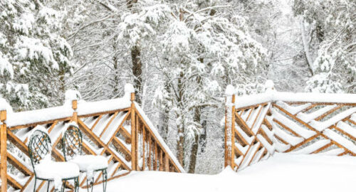 Forest, snow covered house, home wooden deck with two cast iron chairs, snowing, falling snowflakes, snowstorm, storm, trees in backyard, front yard, railing, fence in winter with nobody
