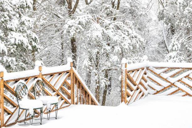 Forest, snow covered house, home wooden deck with two cast iron chairs, snowing, falling snowflakes, snowstorm, storm, trees in backyard, front yard, railing, fence in winter with nobody